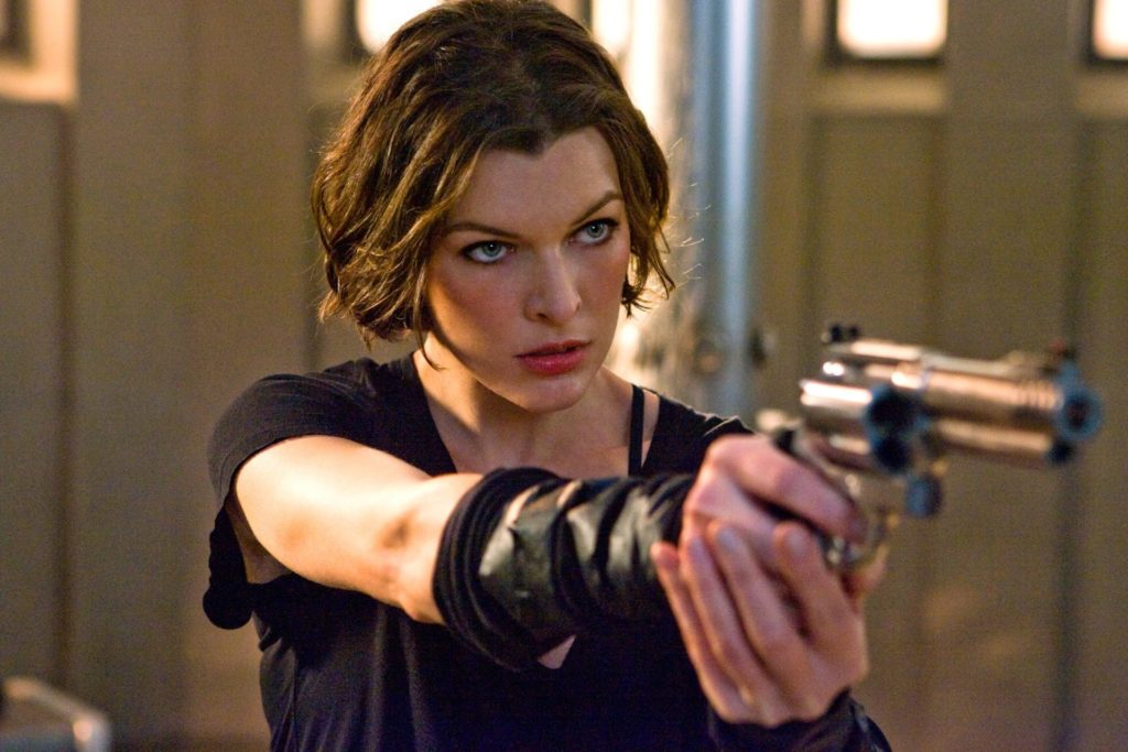 Milla Jovovich em Resident Evil, segurando uma pistola com as duas mãos a frente de seu corpo, mirando para um ponto a frente. Ela olha intensamente para o mesmo ponto.