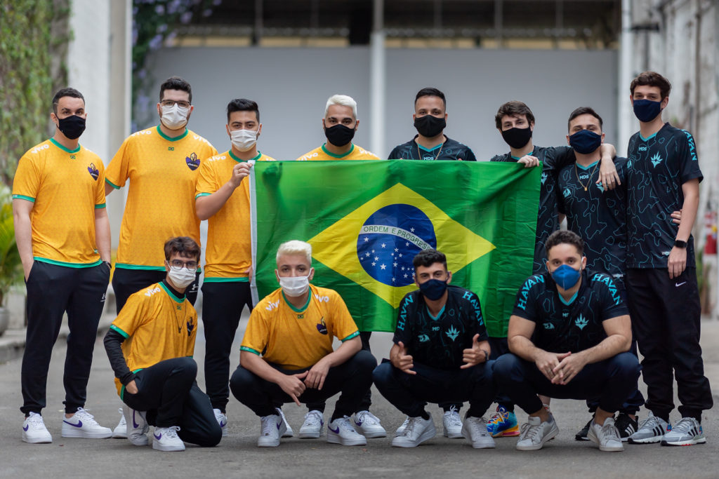 A foto mostra duas lines completas, no ensaio da Riot Games para o Masters Berlim. À esquerda, a equipe da Vivo Keyd, já classificada para o mundial, se concentra em seu uniforme especial, em amarelo com detalhes verdes. À direita está a equipe da Havan Liberty, que jogará o Last Chance Qualifier, em seu uniforme azul escuro. As equipes são unidas pela bandeira do Brasil centralizada, segurada pelos capitães das equipes.